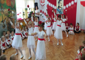 Dziewczynki w białych sukienkach z czerwonymi paskami wykonują układ taneczny z kwiatami.